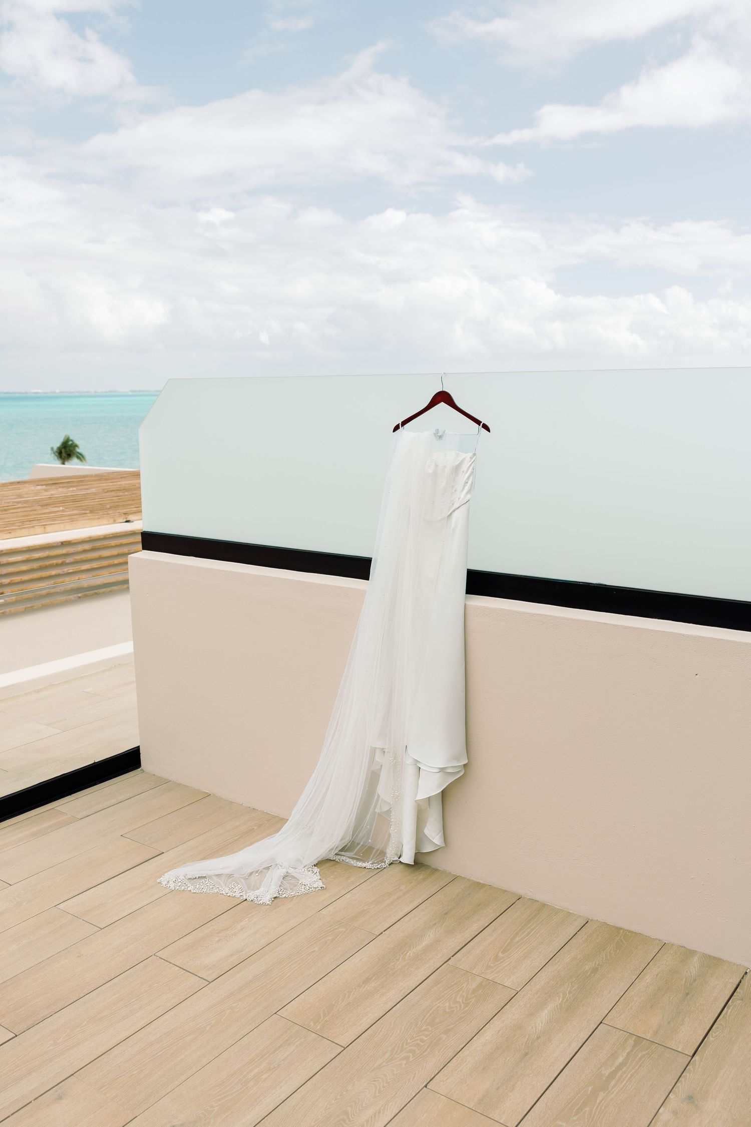 White wedding dress hanging on a balcony hanger with ocean view backdrop on a sunny day.