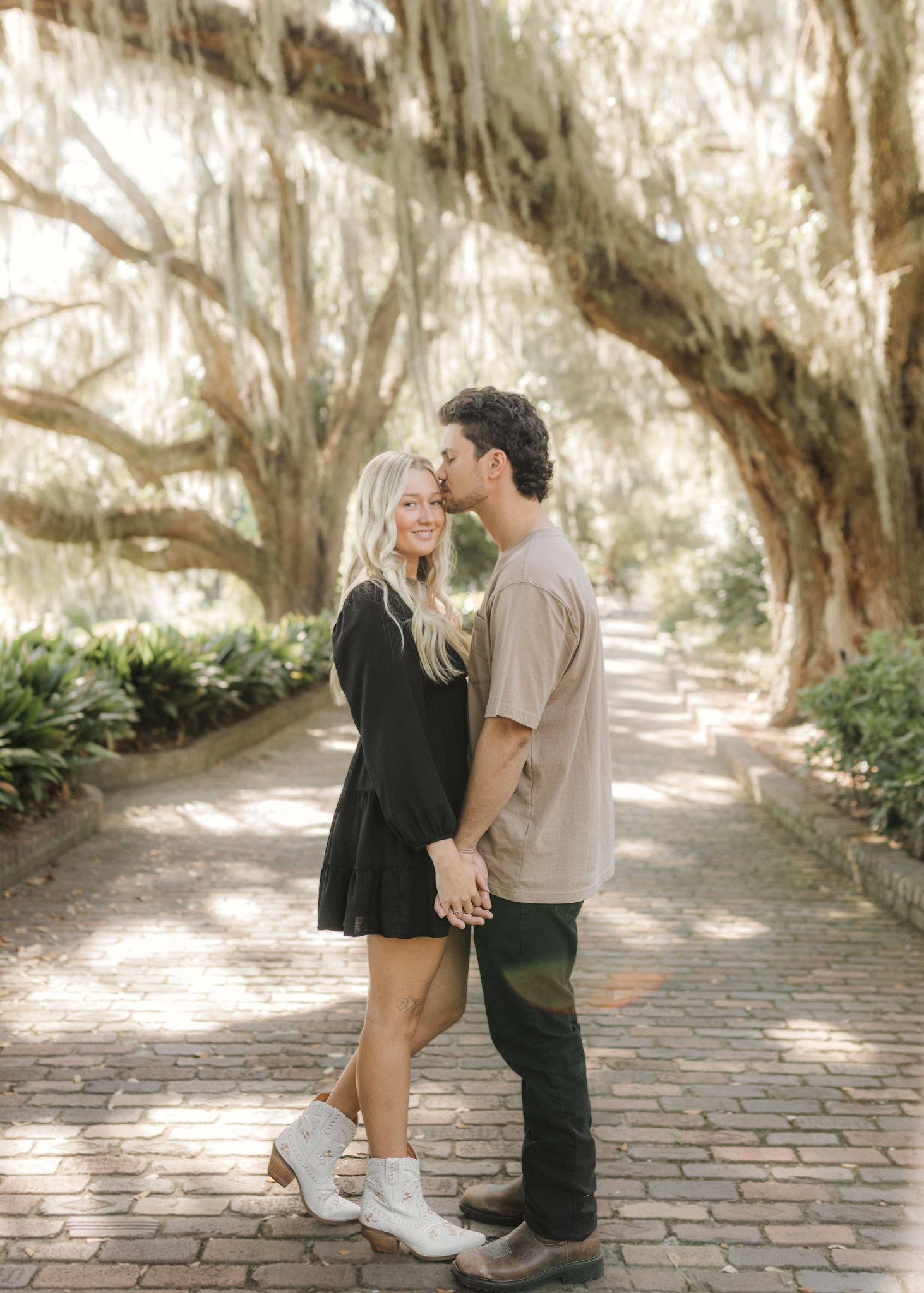 A woman in a black dress hugs a young boy in cowboy boots outdoors among live oak trees on a brick path.