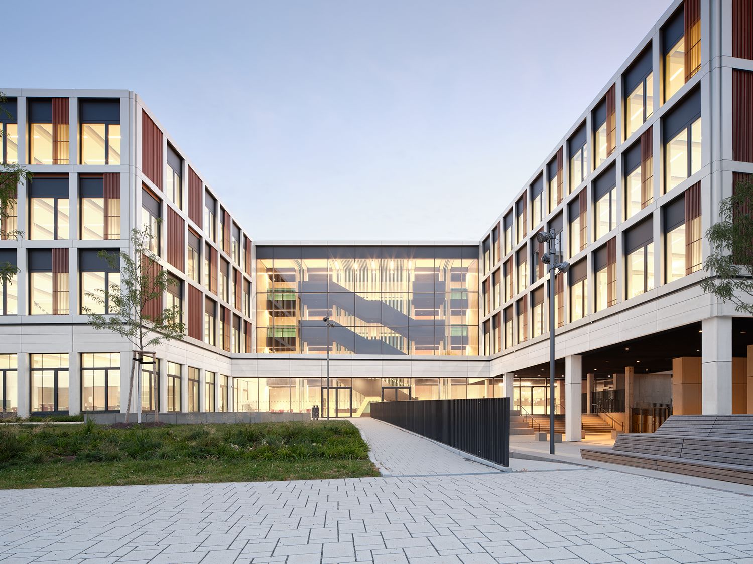 Modern Sporthalle building with a reddish-brown facade and glass entrance, framed by young trees and a quiet street at dusk.