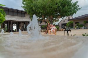 Two people sit together on a bench behind a bubbling fountain, with a large shade tree and rustic buildings in the background.