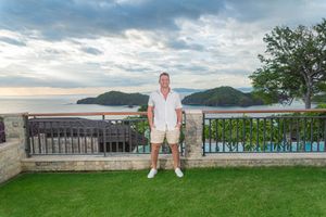 Man in white shirt and khaki shorts stands on a green lawn by a stone terrace railing overlooking a scenic ocean bay with forested islands.