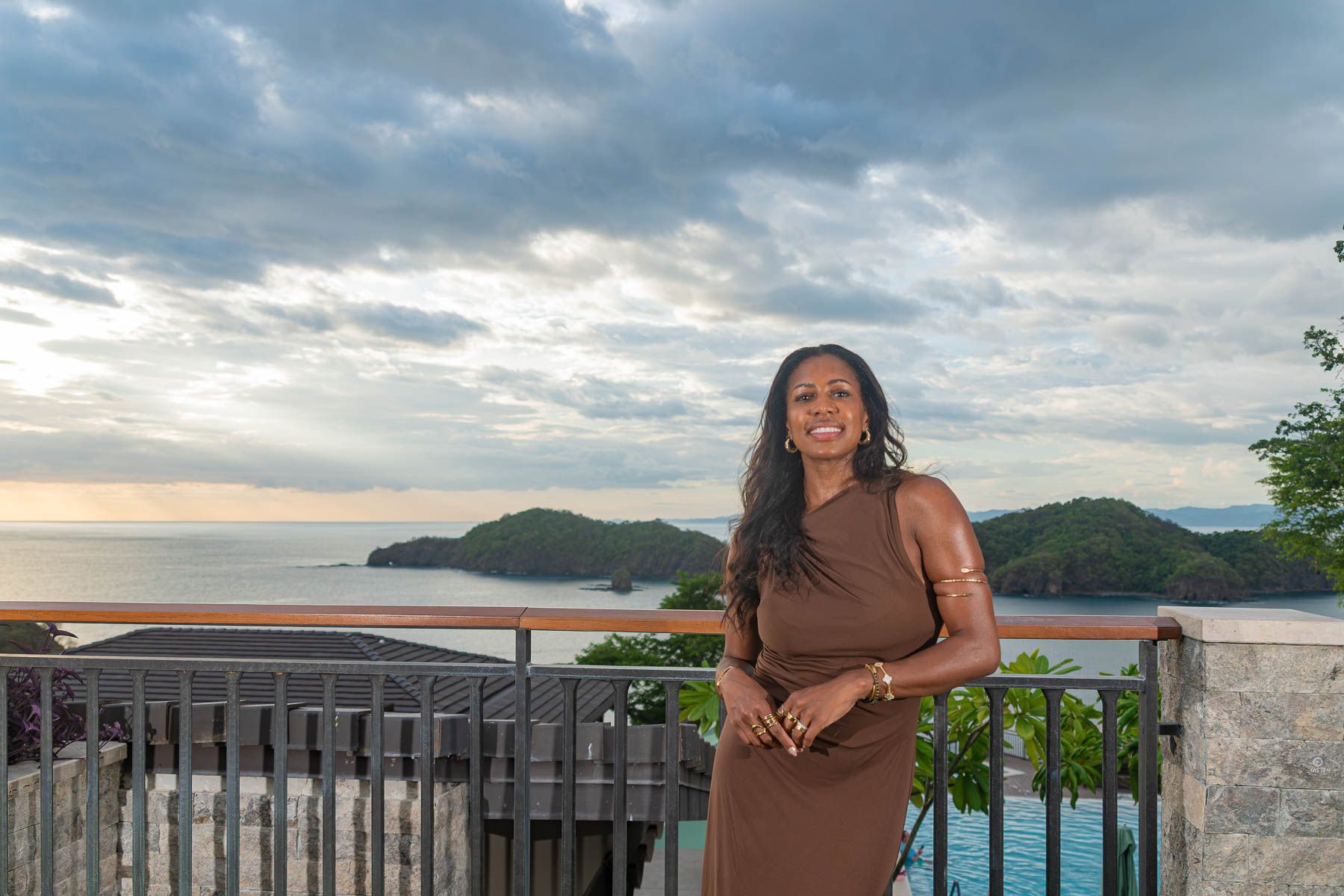 Woman in a brown dress stands on a hillside terrace railing overlooking the ocean and green islands at sunset under a cloudy sky.
