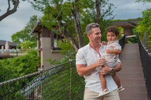 Smiling man in a white shirt holds a toddler on a wooden walkway with a railing, surrounded by lush green trees at a tropical resort.