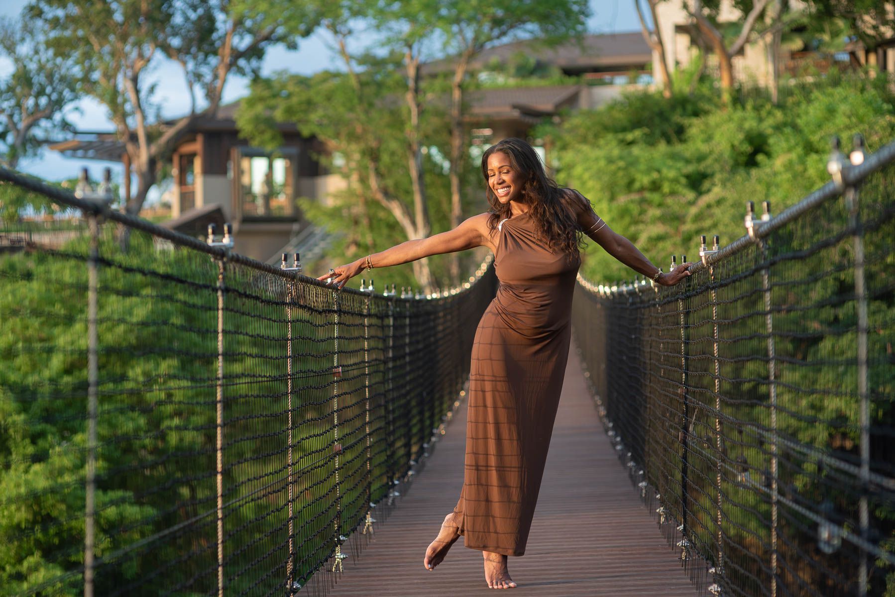 Smiling woman in a brown dress walks barefoot across a wooden suspension bridge with rope netting railings, surrounded by lush green trees.