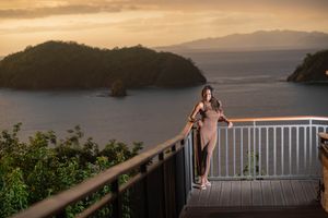 Woman in a sheer wrap stands on a wooden balcony overlooking forested islands and calm ocean waters at golden sunset.