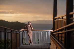 Woman in a fitted tan dress leans on a hillside balcony railing, gazing at a golden sunset over distant mountains and the sea.
