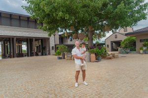Man in white shirt and shorts holding a toddler on a brick courtyard beside a large shade tree and rustic stone building.