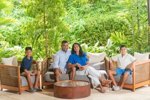Family of four relaxing together on cushioned wicker patio seating around a round copper coffee table surrounded by lush tropical greenery.