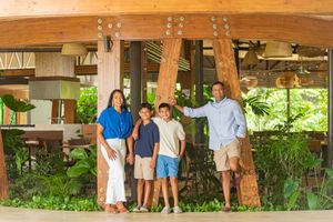 Smiling family of four poses together beneath wooden beams in an open-air tropical resort lobby surrounded by lush green plants.