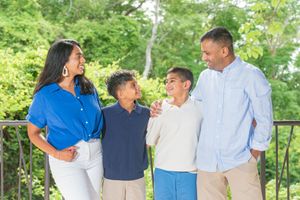 Smiling family of four in coordinated blue and white outfits standing together on a balcony with lush green trees in the background.
