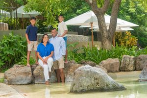 Family of four posing together beside a shallow pool with large boulders, lush tropical plants, and shade umbrellas at a resort.