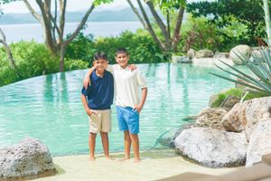 Two smiling boys stand with arms around each other beside a turquoise infinity pool surrounded by rocks, tropical plants and trees.