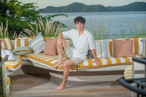 Young man in a white shirt and shorts sits barefoot on a striped outdoor daybed with cushions overlooking a calm bay and green hills.