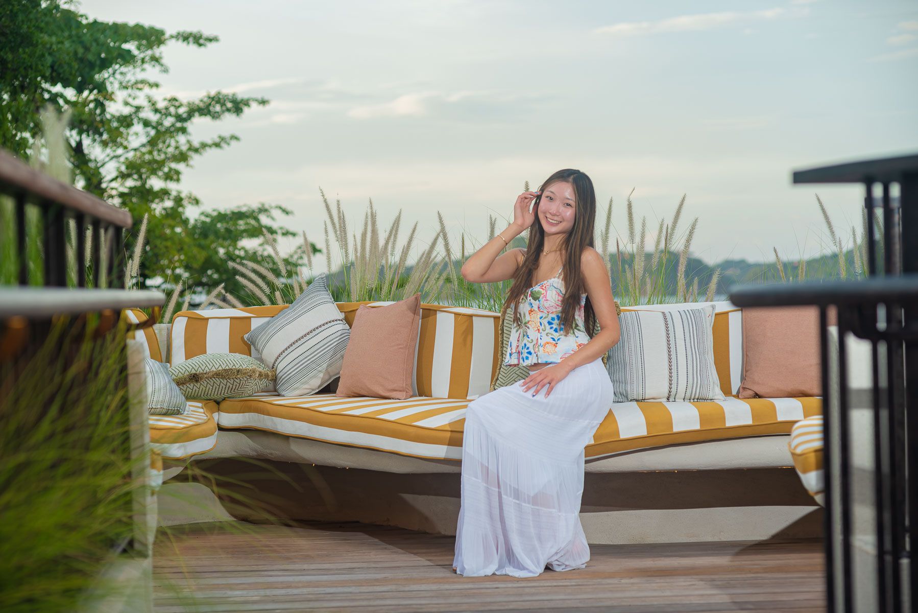 Smiling woman in a white maxi skirt and floral top sits on a striped outdoor daybed with orange pillows on a terrace with tropical plants.