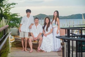 Family of four in white and neutral outfits posing barefoot on a wooden deck with a scenic bay and green hills in the background.