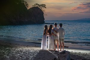 Four people stand arm in arm on a quiet beach at sunset, gazing at calm ocean waters and a rocky headland under a glowing pink sky.