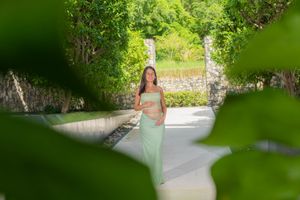 Pregnant woman in a flowing mint green dress cradles her belly while standing on a sunlit garden path framed by lush green leaves.