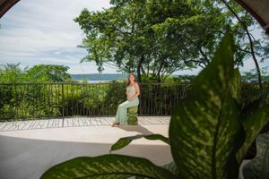Woman in a long green dress sits on a stone stool on a sunny terrace overlooking lush tropical trees and the distant ocean.