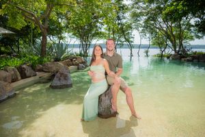 Smiling couple sits together on a rock beside a clear tropical lagoon pool surrounded by lush green trees at a scenic resort.