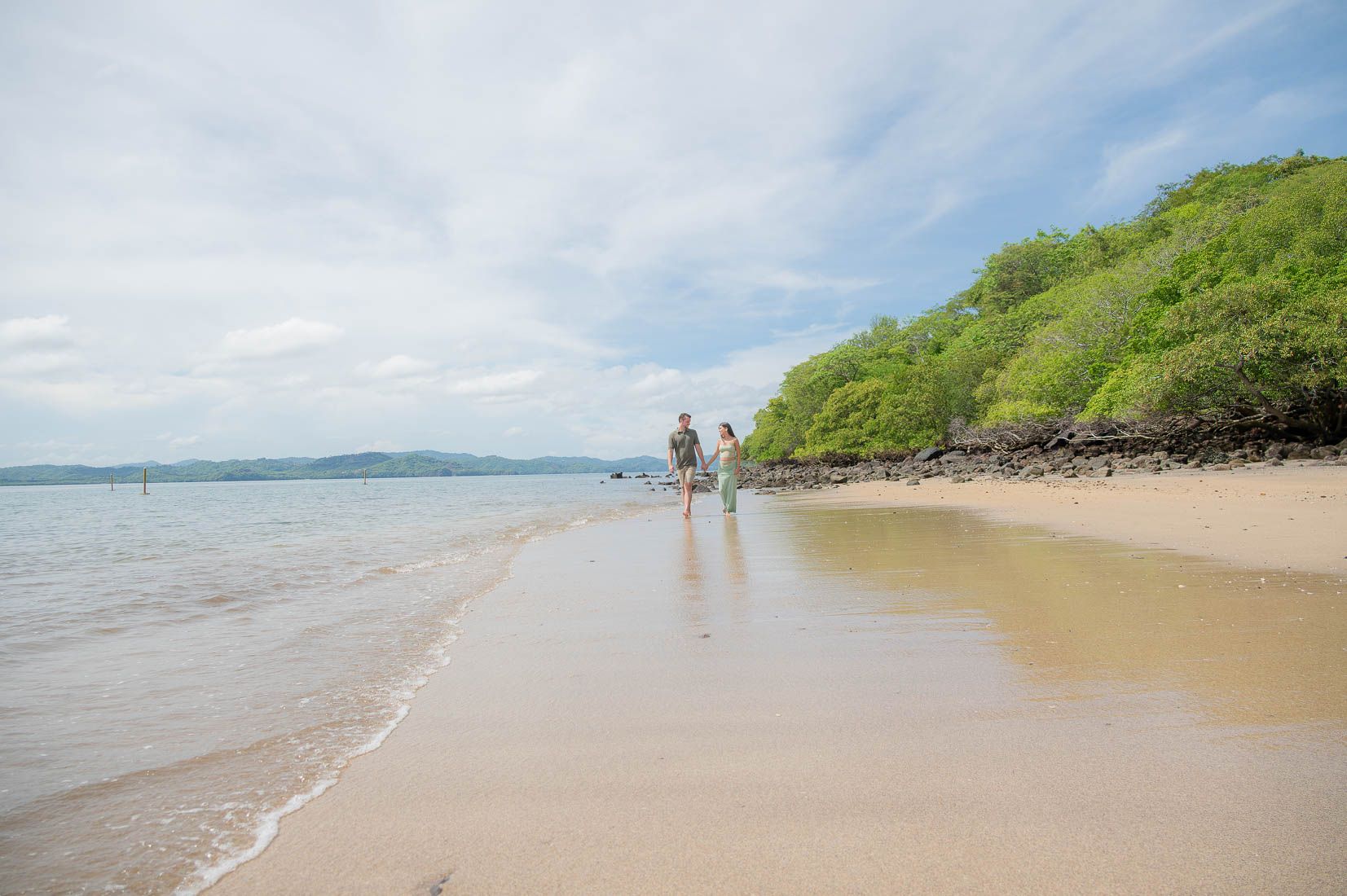 Two people walk along a wet sandy beach beside calm turquoise water and a lush green hillside under a partly cloudy sky.