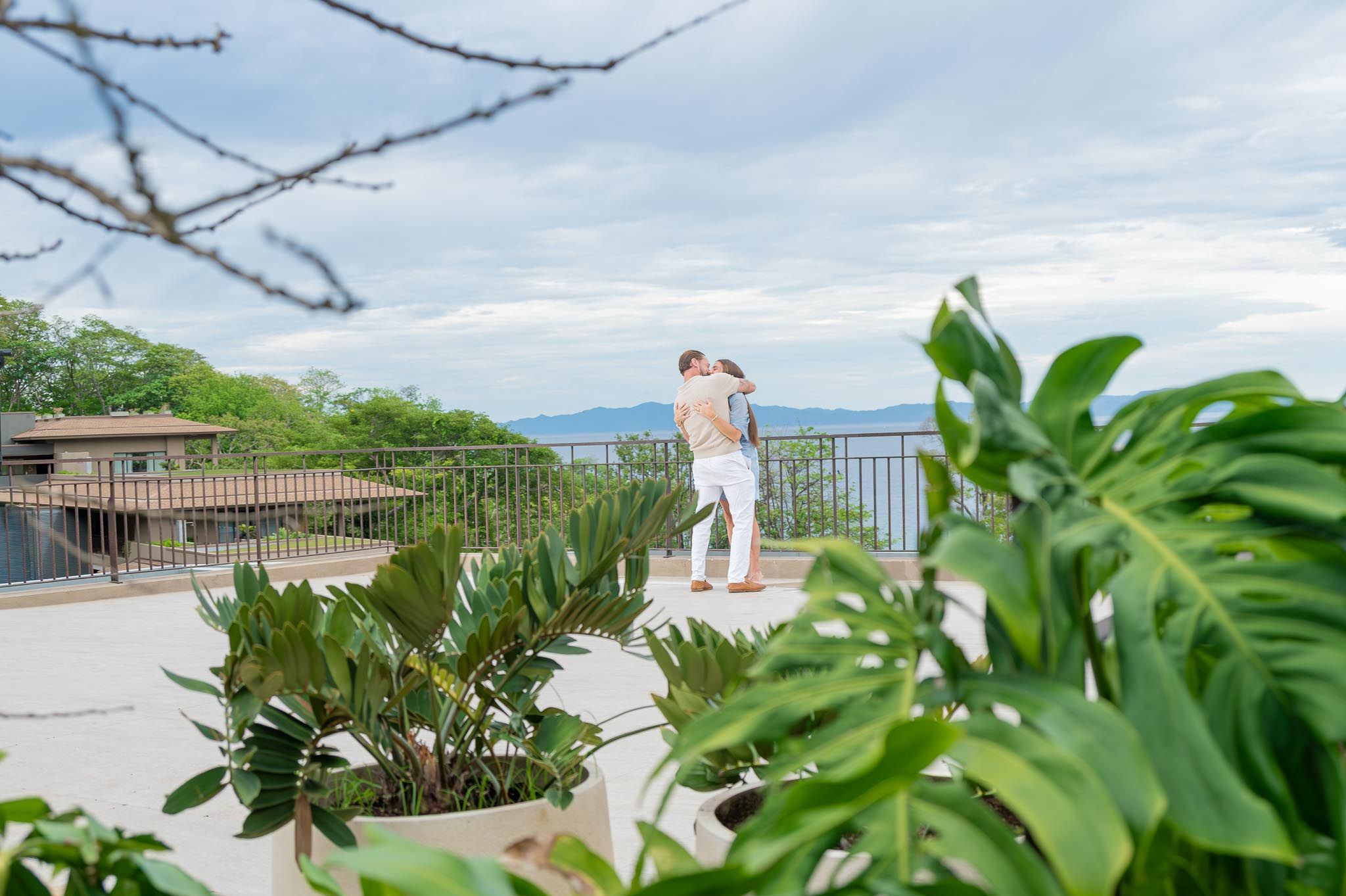 Couple embracing on a sunny terrace framed by lush green tropical plants, with a villa, railing and distant mountain view in the background.