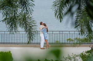 Couple embracing on a seaside promenade beside a railing, framed by tropical palm leaves with calm blue water behind them.