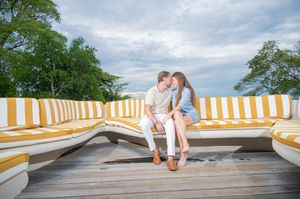 Couple sitting close together on a yellow and white striped curved outdoor bench on a wooden deck, with trees and a cloudy sky behind.