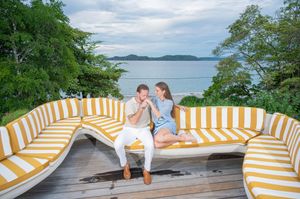 Romantic couple sitting close on a curved yellow and white striped outdoor sofa on a wooden deck overlooking a calm bay and green hills.