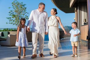 Smiling family of four in light summer outfits walking hand in hand along a sunny resort terrace with ocean views.