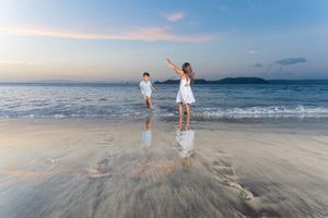 Two children in white outfits run and play along the wet sand of a calm beach at sunset, with gentle waves and distant hills behind.
