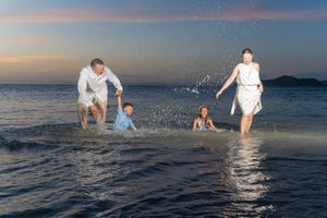 Family of four playfully splashing in shallow ocean water at sunset, with parents and two children laughing against a colorful twilight sky.