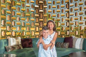 Two smiling children embrace in front of a teal green sofa with patterned pillows and a decorative golden geometric wall.