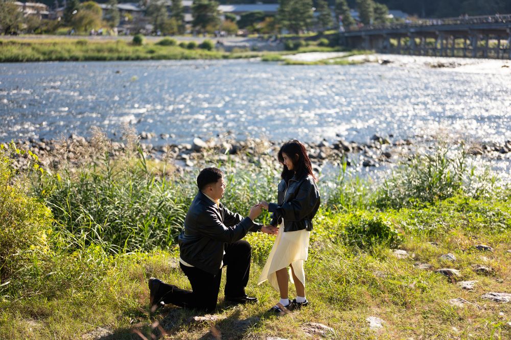 Man kneeling on one knee proposes to a smiling woman on a grassy riverbank with sparkling water and a bridge in the background.