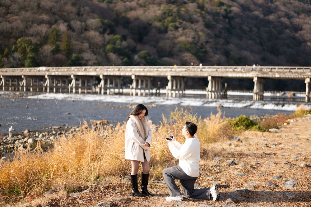 Man kneeling on one knee proposing to a woman in a white coat on a grassy riverbank with a long bridge and autumn hills behind them.