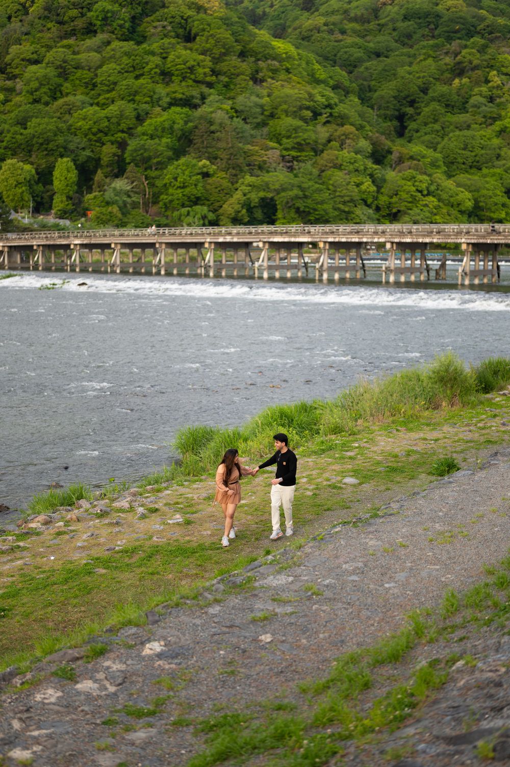 Couple holding hands and walking along a grassy riverbank with a long wooden bridge and lush green forested hills in the background.