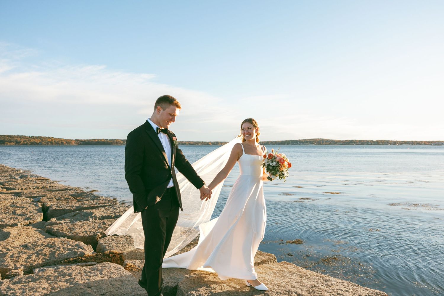 Bride in a white gown and groom in a black tuxedo hold hands while walking along a rocky shoreline at sunset on their wedding day.