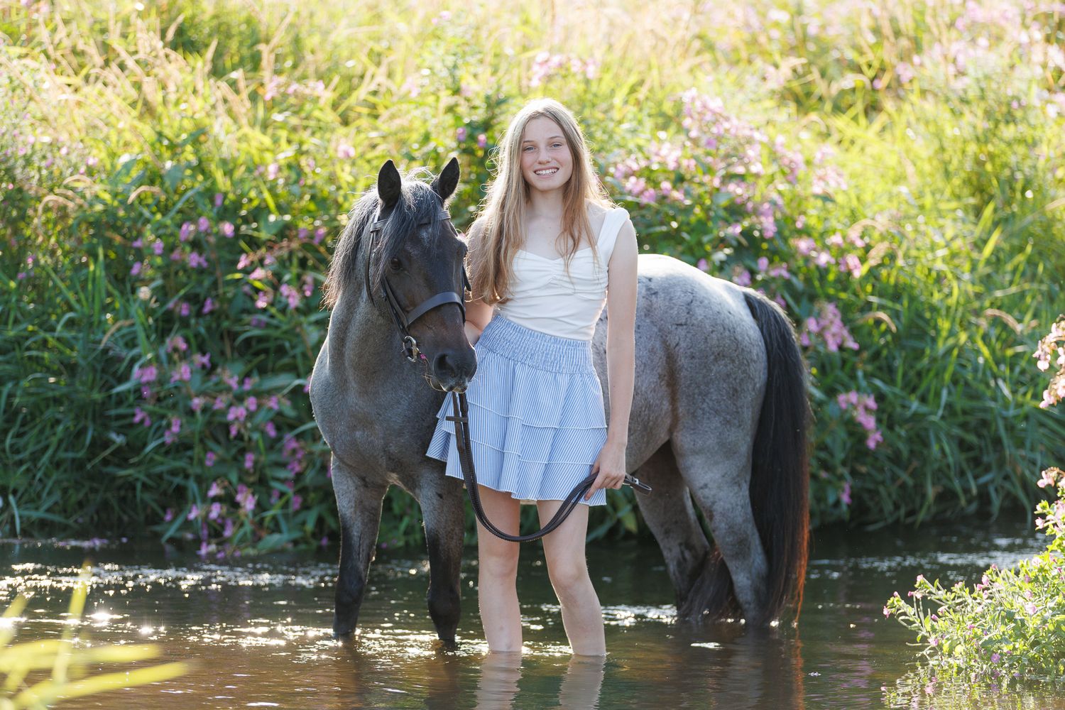 Woman walking two chestnut horses down a sunlit gravel path lined with green trees at a horse farm.