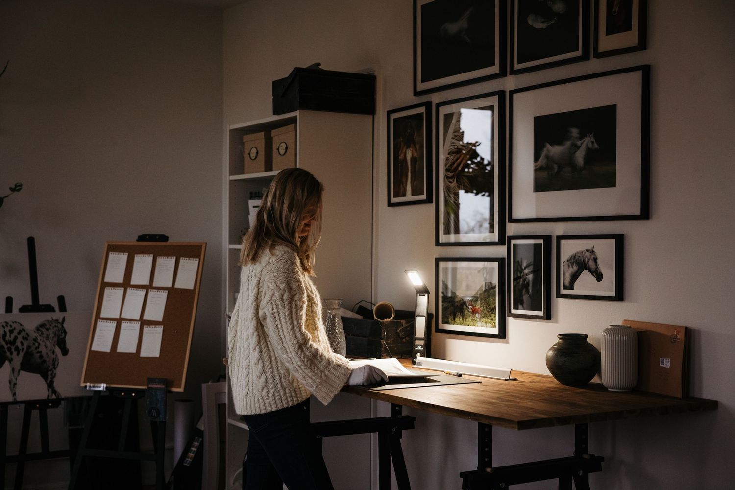 Woman in white sweater working at a wooden desk by candlelight in a dark art studio with framed pictures on the wall.