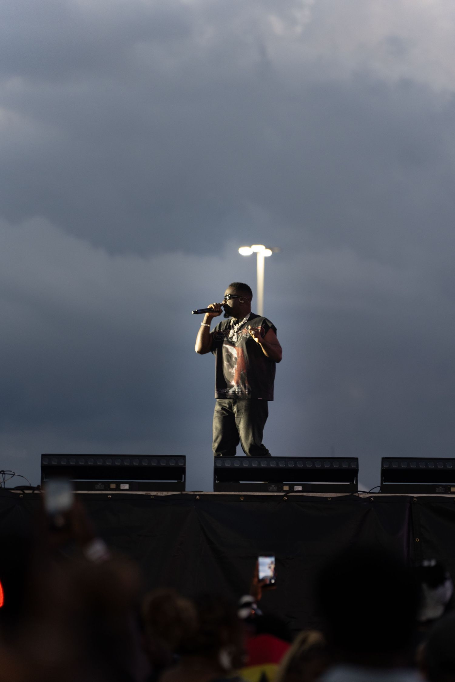 Male performer singing on outdoor stage with dramatic cloudy sky and spotlight behind him at a live concert.