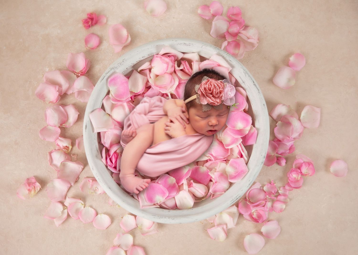 Newborn baby girl sleeping in a white bowl surrounded by pink rose petals, wearing a floral headband.
