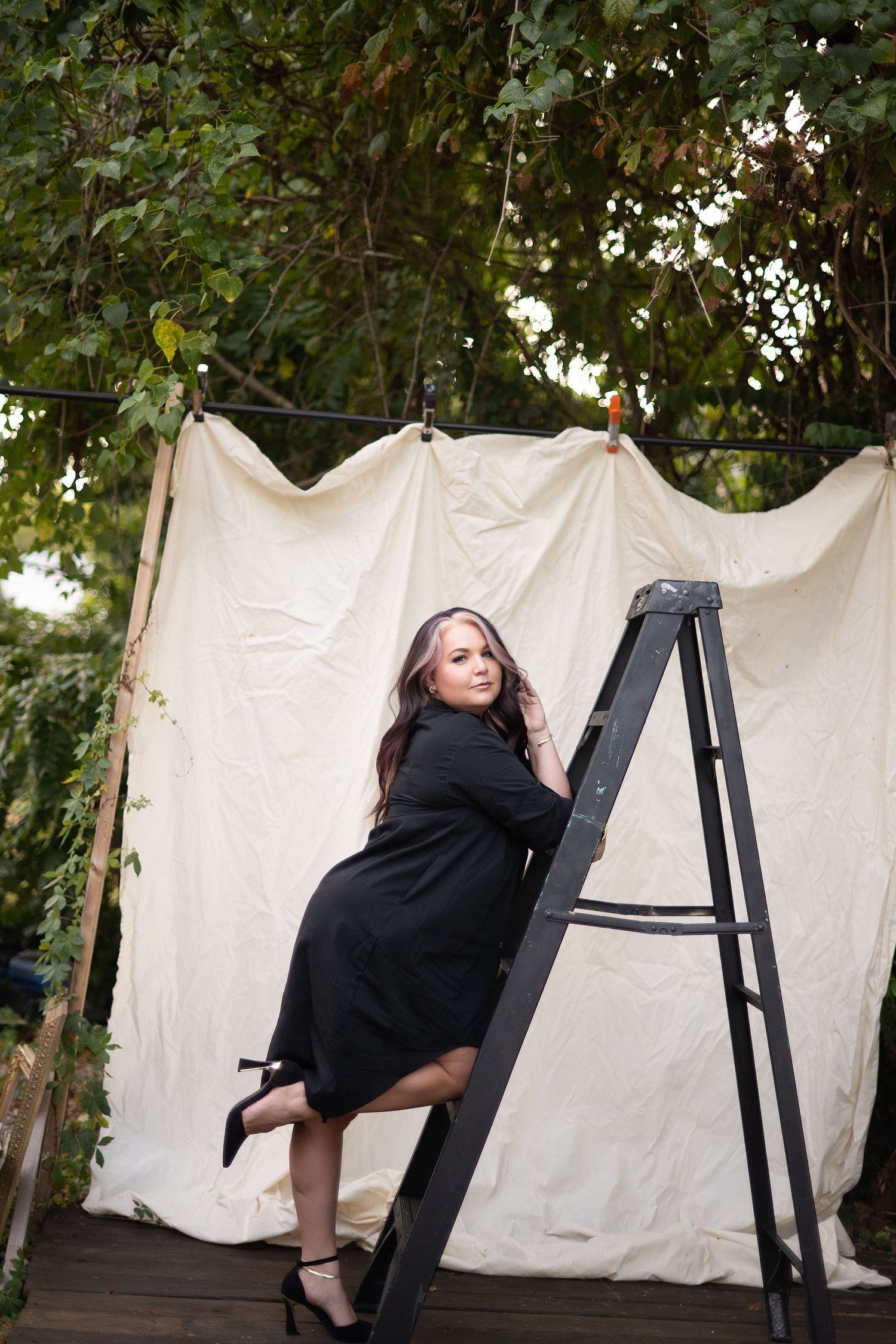 Woman in black dress poses on a ladder outdoors against a white fabric backdrop surrounded by lush green trees.