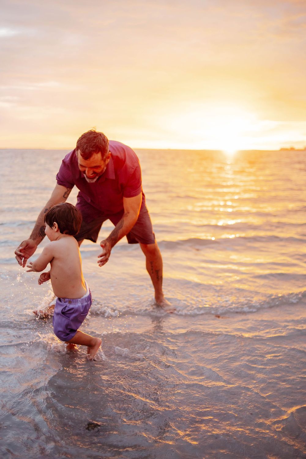 Smiling father in a maroon shirt helps his toddler in purple swim trunks walk through shallow ocean water at a golden sunset beach.