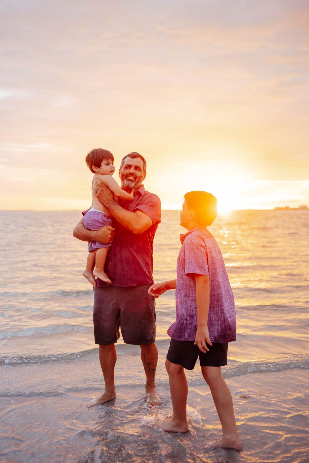 A father holding a toddler stands barefoot in shallow ocean water beside a boy as the golden sunset glows over the beach.