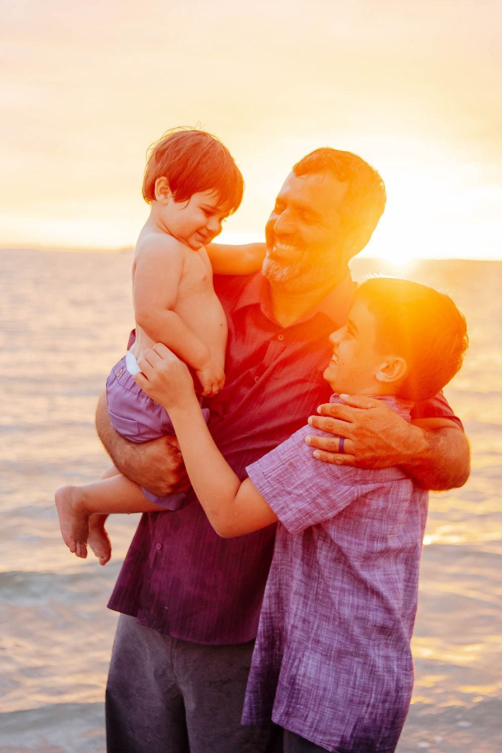 A smiling man in a maroon shirt holds a young red-haired child while hugging an older boy on a beach at golden sunset.