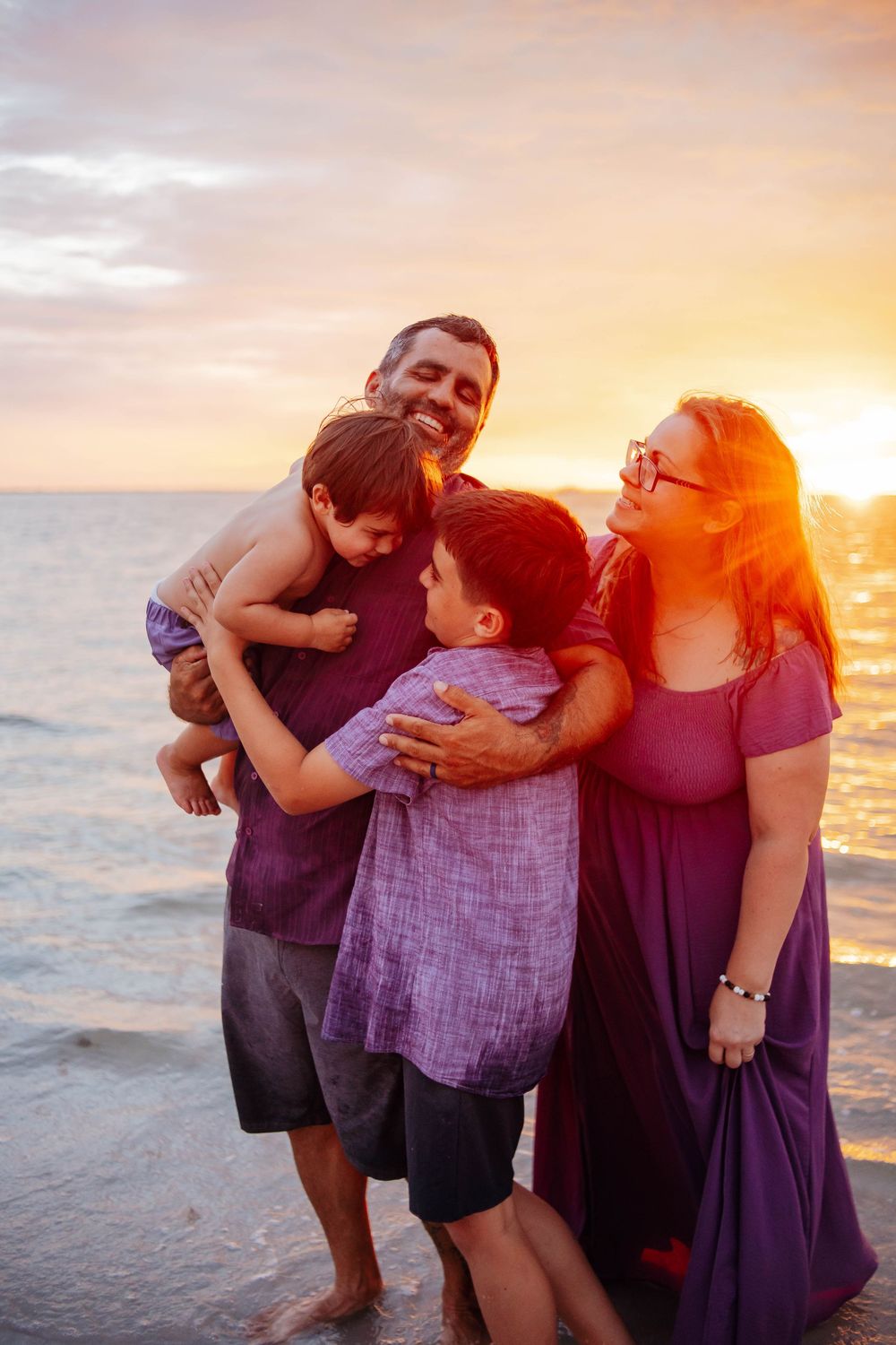 A smiling family of four hugs on the beach at sunset, with two boys embraced by their parents beside the glowing ocean.