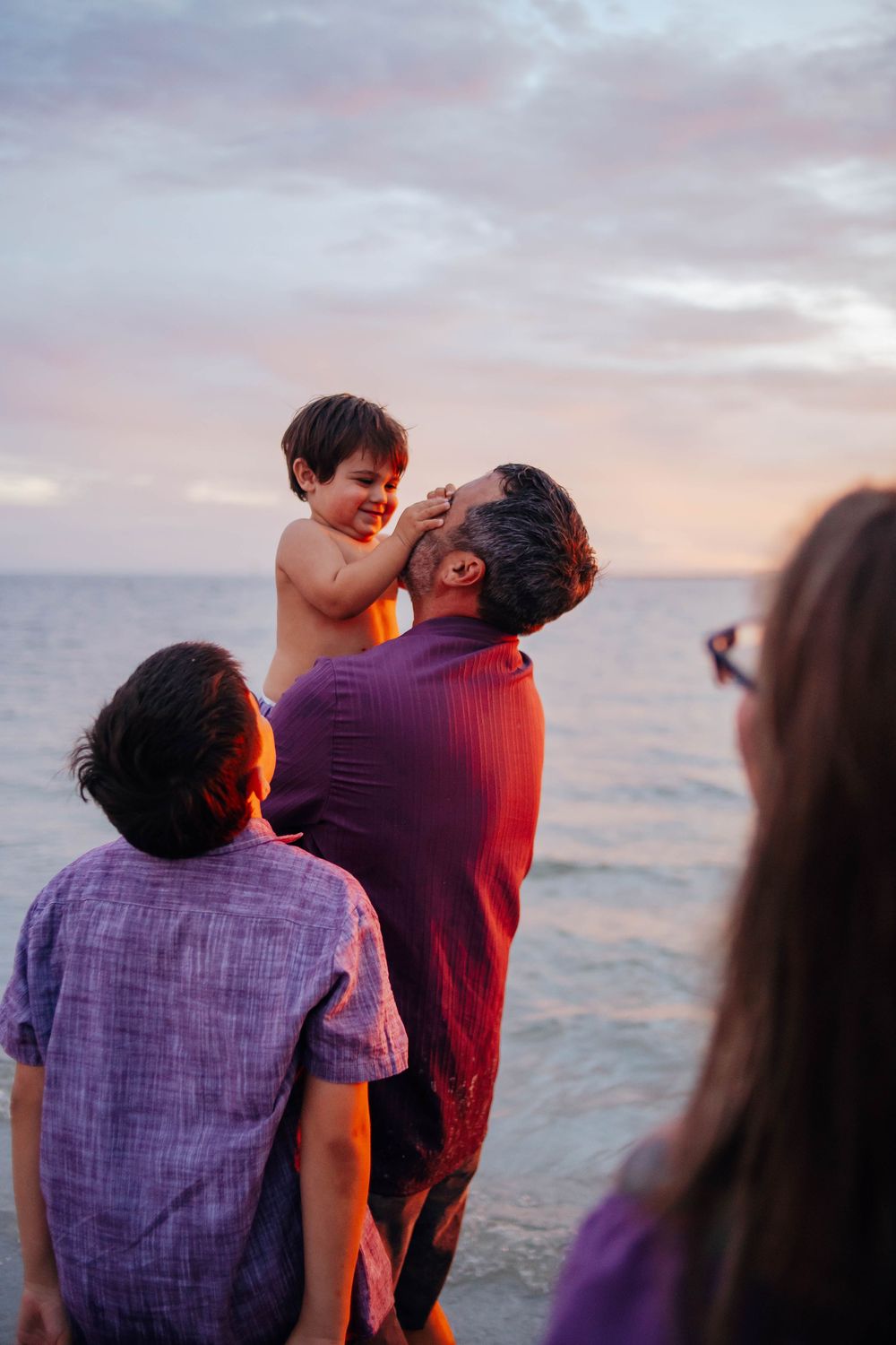 Family on a beach at sunset as a smiling father lifts a laughing young boy while a child and woman watch the calm water.