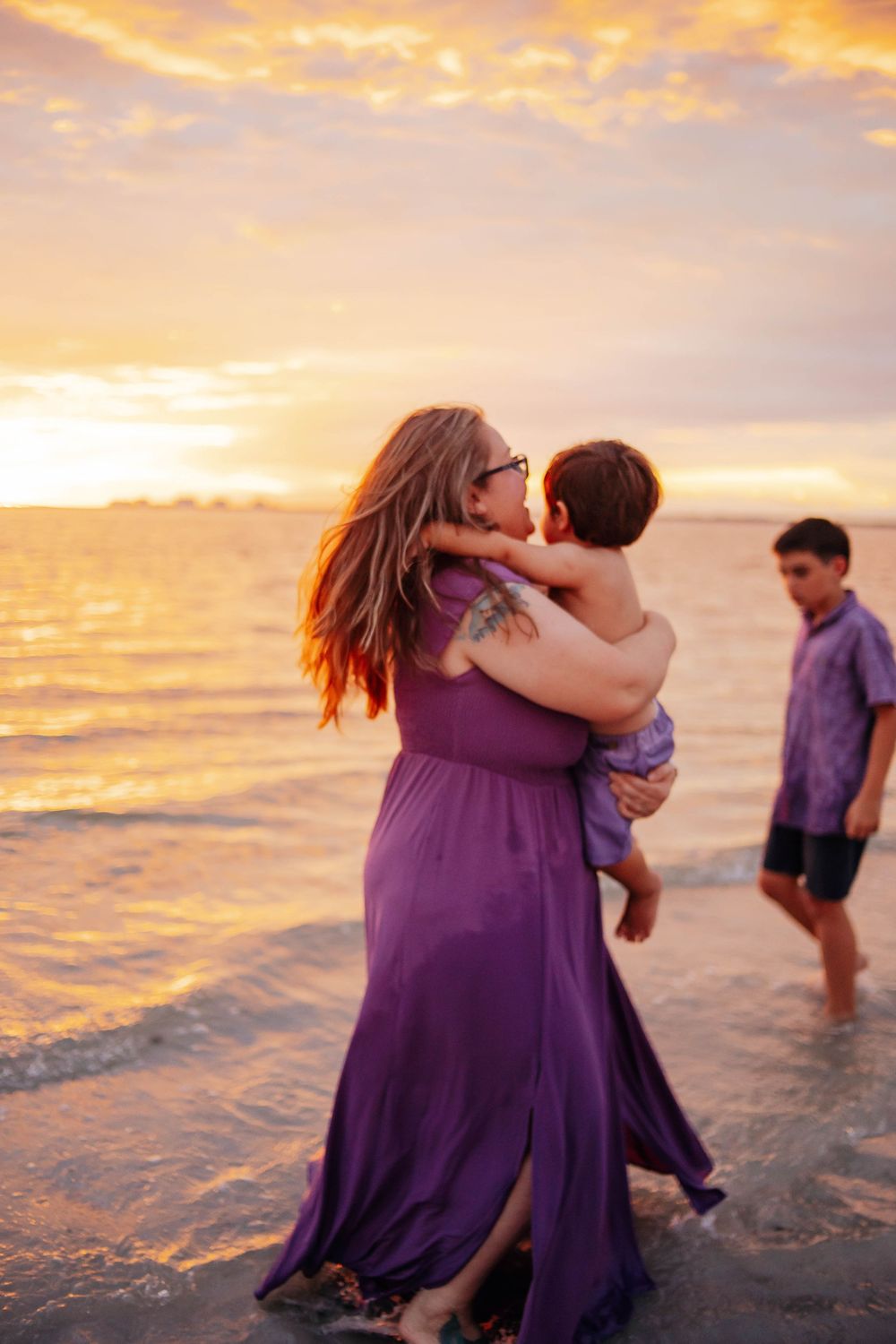 A woman in a purple dress holds a toddler in the shallow ocean water at sunset while an older boy walks nearby.