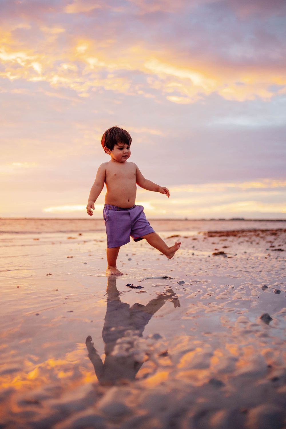 Toddler in purple shorts playing on a wet sandy beach at sunset with a golden pink sky and reflection in the shallow water.