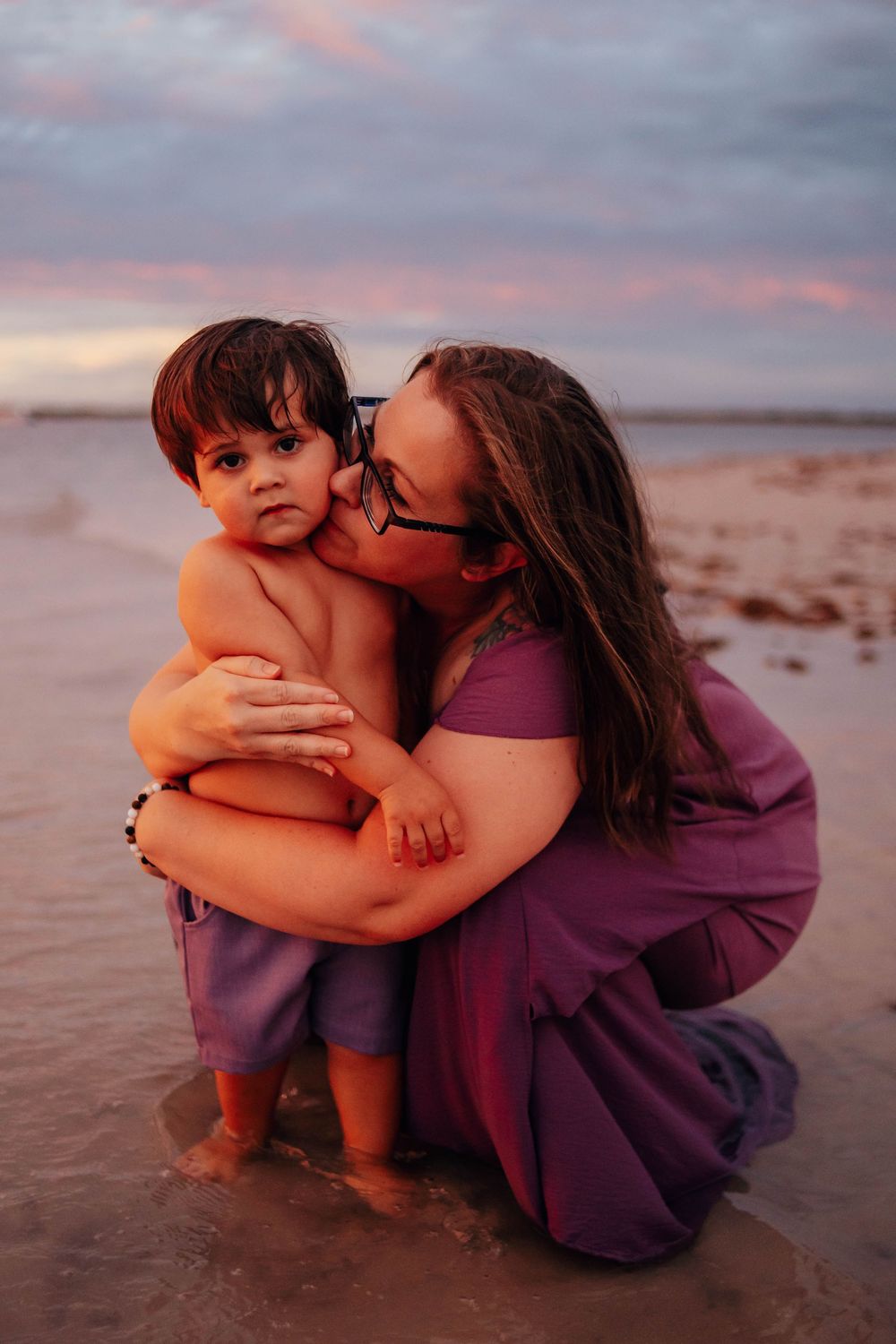 A woman in a purple dress kneels in shallow beach water at sunset, hugging and kissing a smiling shirtless toddler on the cheek.