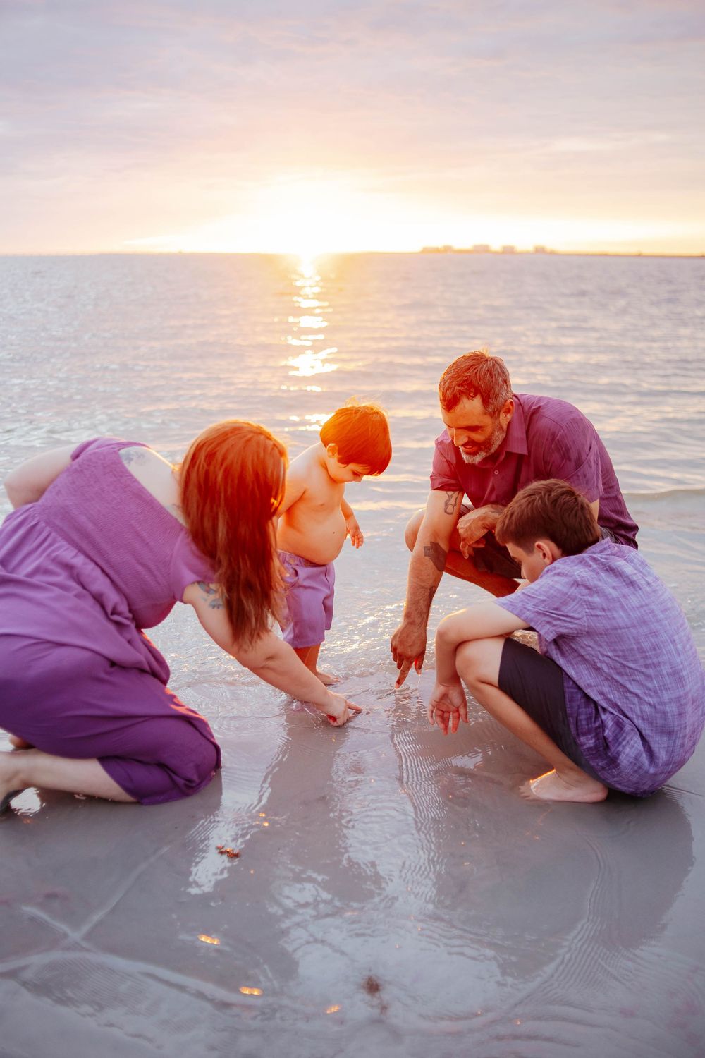 A family of four kneels together on a wet sandy beach at sunset, drawing in the sand beside the calm, golden ocean.
